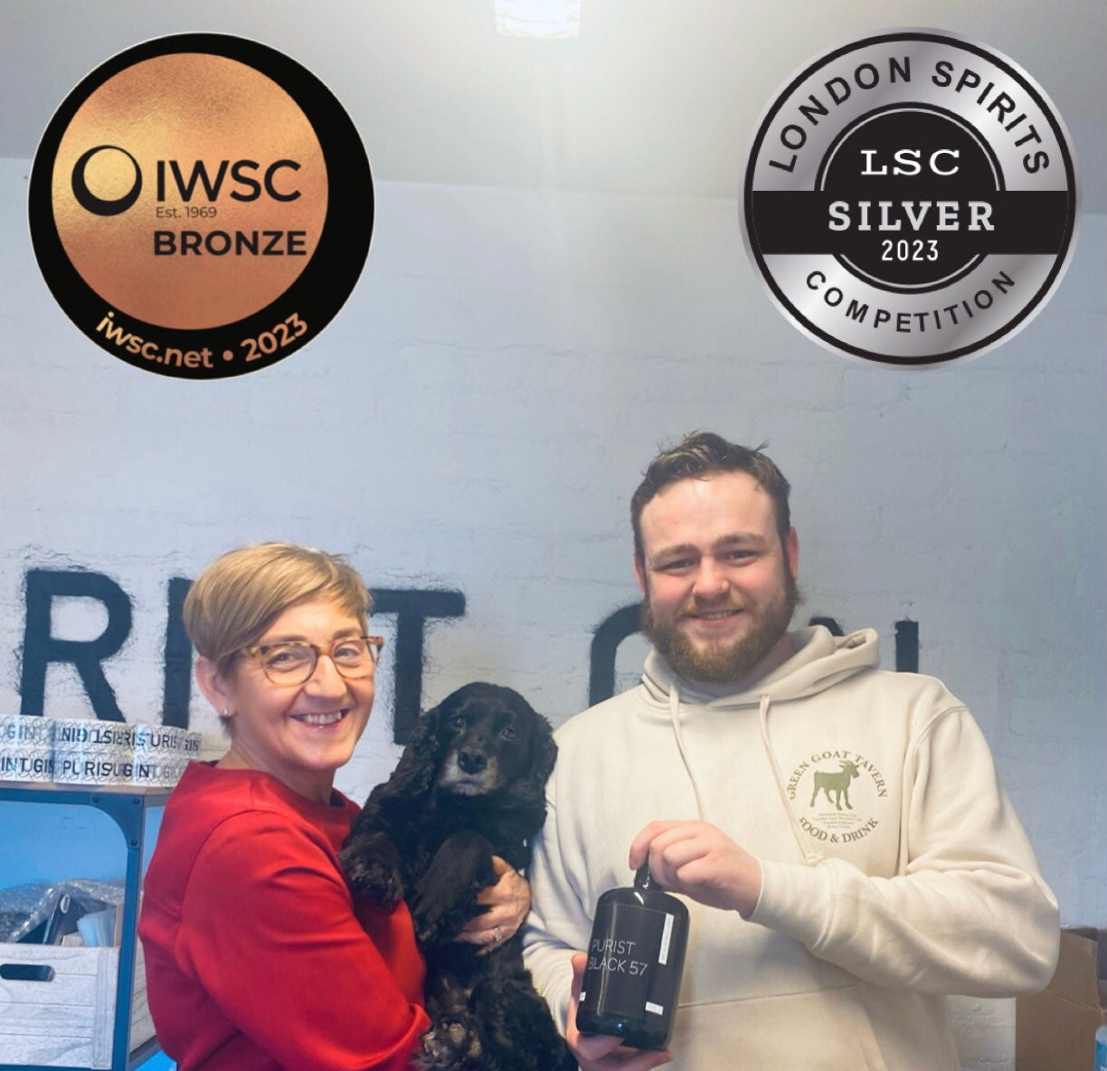 Colette, Charlie the Dog, and Bruce holding a bottle of Purist Black 57 gin with two medals at the top of the image. One is the bronze medal with IWSC and the other is a silver medal with the London Spirit Competition. 
